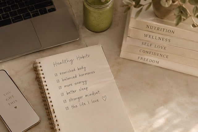Workspace with spiral notebook listing healthy habits, laptop, green smoothie, and wellness note cards on beige desk