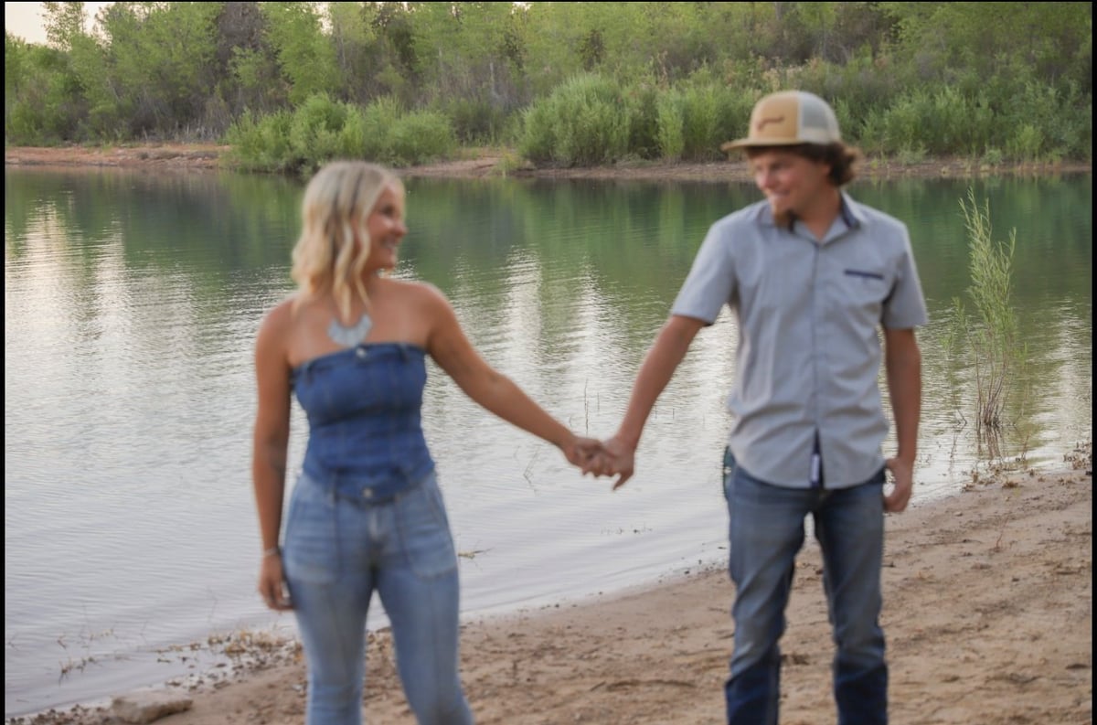 A couple holding hands on a sandy riverbank with forested hills and calm water in the background